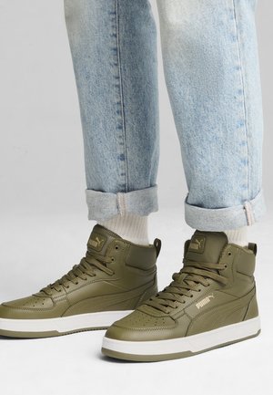 Persona che indossa jeans azzurri arrotolati, calzini bianchi e sneaker alte Puma color verde oliva con suole bianche.