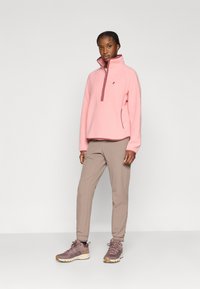 Ανοιχτό ροζ φλις πουλόβερ με ψηλό γιακά και φερμουάρ μέχρι τη μέση, συνδυασμένο με μπεζ παντελόνι και μοβ παπούτσια πεζοπορίας. Υλικό με υφή και απλός σχεδιασμός.