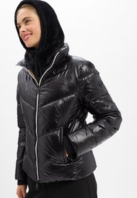 Femme portant une doudoune noire brillante avec une capuche en fausse fourrure noire, les mains dans les poches, souriante, sur fond blanc uni.