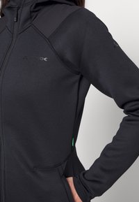 Svart huva jacka med texturerad tyg, med dragkedjestängning, sidofickor och grön accent vid nederkanten. Inkluderar logotypdetalj på bröstet.