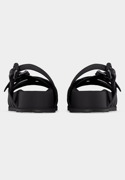 Ζευγάρι μαύρων φλατ σανδαλιών με πολλαπλές λωρίδες και μεταλλικές αγκράφες, από πίσω σε απλό φωτεινό φόντο.