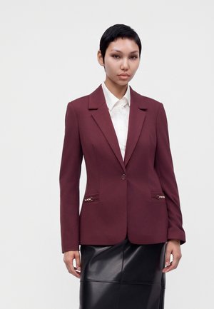 Mujer con cabello corto y oscuro, vistiendo un blazer burdeos sobre una camisa blanca y una falda de cuero negra contra un fondo claro y liso.