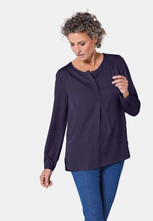 Blusa a maniche lunghe blu navy con patta di bottoni, tessuto morbido, dettagli plissettati sulle spalle e vestibilità rilassata. Indossata con jeans blu.
