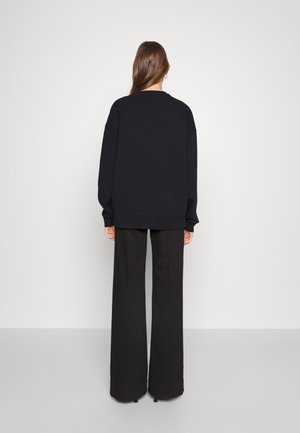 Femme aux longs cheveux bruns portant un sweat-shirt noir oversize et un pantalon large noir, debout face à un mur blanc uni.