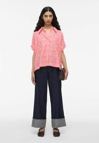 Roze gemêleerd overhemd met korte mouwen, gecombineerd met brede donkere denim jeans met een omgeslagen zoom. Houdt een kleine bruine tas vast.