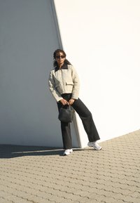 Giacca trapuntata beige chiaro con colletto nero, pantaloni neri a gamba larga, sneakers bianche e una borsa nera su uno sfondo luminoso.