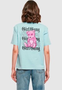 Gaiši zils kokvilnas T-krekls ar lielu rozā lāča grafiku un "Girl Gang" tekstu trešajā pusē, rakstītu drosmīgā un stilizētā fontā.