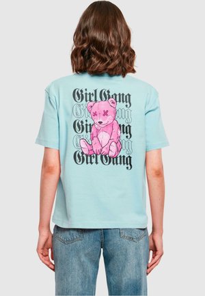 Lyseblå bomulds T-shirt med et stort pink teddy bear grafik og teksten "Girl Gang" i en fed, stiliseret skrifttype på ryggen.