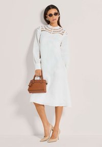 Frau in einem weißen Midi-Kleid mit langen Ärmeln und Ausschnitten, beigen High Heels, dunkler Sonnenbrille und einer kleinen braunen Handtasche.