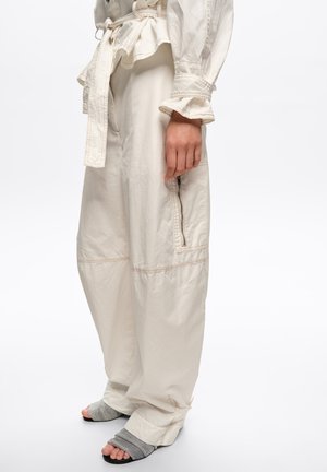 Persona con pantalones sueltos de color crema con cremallera lateral y cinturón en la cintura, combinados con sandalias grises de dedo abiertas.