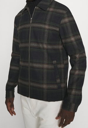 Uomo che indossa una giacca a zip a quadri scuri sopra un dolcevita nero e pantaloni bianchi, in piedi contro uno sfondo neutro.