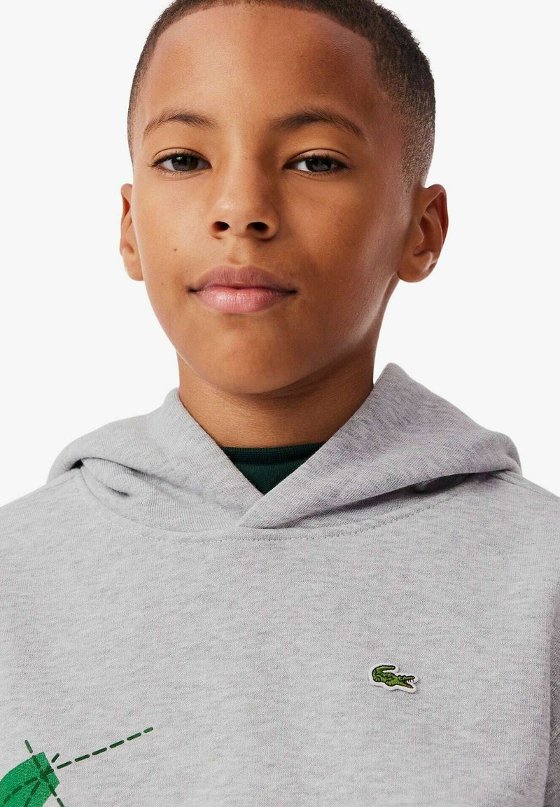 Grijze hoodie met een kangoeroezak, voorzien van een groene geborduurde krokodillogo op de borst, gedragen over een donkergroene shirt.