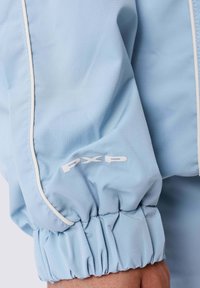 Manica di giacca azzurra chiara con polsini elasticizzati. Presenta piping bianco e un logo ricamato in bianco. Tessuto morbido e leggero.