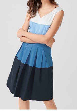 Vrouw met een mouwloze jurk in kleurvlakken met witte, lichtblauwe en donkerblauwe horizontale secties, staand met gekruiste armen.