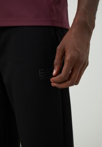 Podrobno posnetek roke osebe, ki počiva blizu črnih hlač z diskretnim logotipom "EA7 Emporio Armani", oblečena v temno rjav vrh.