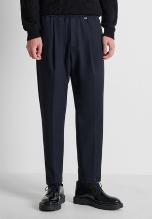 Hombre con pantalones negros hechos a medida, suéter de punto negro y zapatos negros brillantes de cordones, de pie sobre suelo gris.