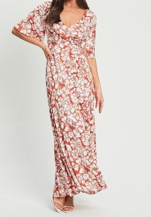 Vrouw draagt een lange, vloeiende jurk met witte en roestkleurige bloemenpatronen, een diepe V-hals, ellebooglange mouwen en witte open-toe hakken.