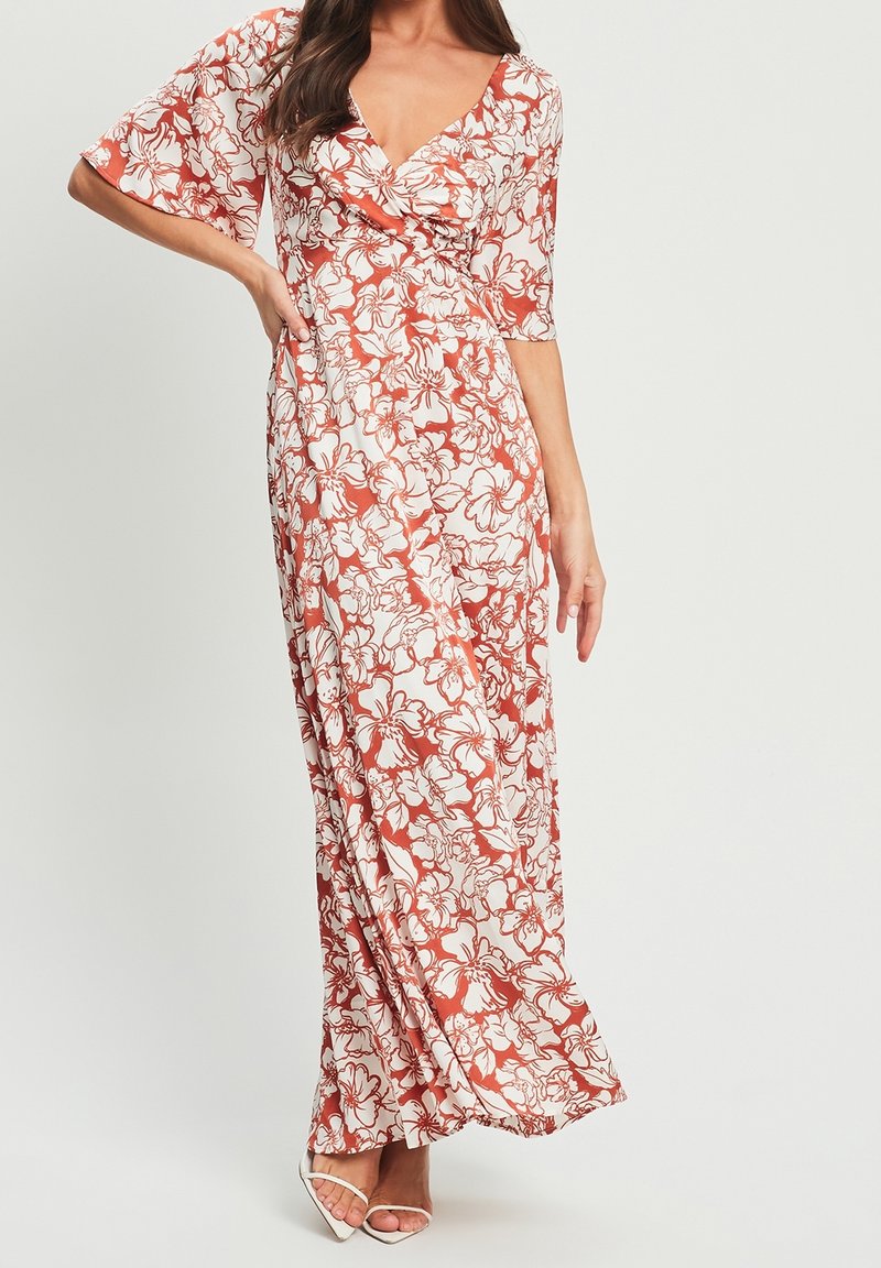 Femme portant une longue robe fluide avec des motifs floraux blancs et rouille, un décolleté en V profond, des manches jusqu'aux coudes et des talons ouverts blancs.
