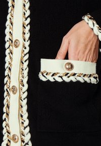 Cárdigan negro de punto con acentos blancos, botones dorados y un trenzado a lo largo de la tapeta de botones y los bolsillos, con un patrón texturizado.