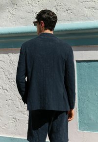 Marineblå tekstureret blazer med lange ærmer, båret over matchende shorts, med et subtilt mønster og en strømlinet silhuet.