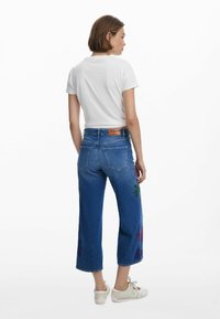 Blå denimjeans med vida ben, prydda med färgglad blommig brodyr på sidan. Matchas med en figurnära vit t-shirt och ljusa skor.