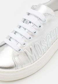 Silver metallic sneaker med vita snören, med präglad "Moschino"-logotyp på sidan och en vit gummisula.