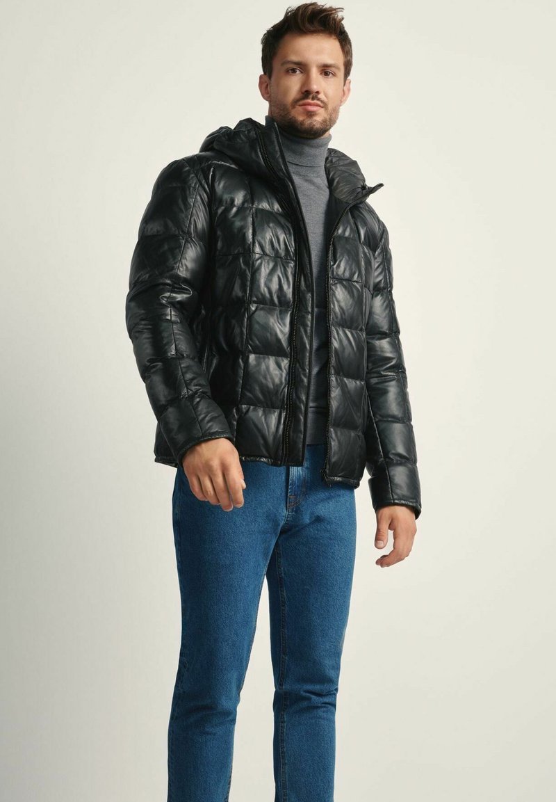 Czarna ocieplana kurtka z kapturem, z zamkiem błyskawicznym i pikowanym wzorem. Nosi ją w połączeniu z szarym golfem i niebieskimi dżinsami.