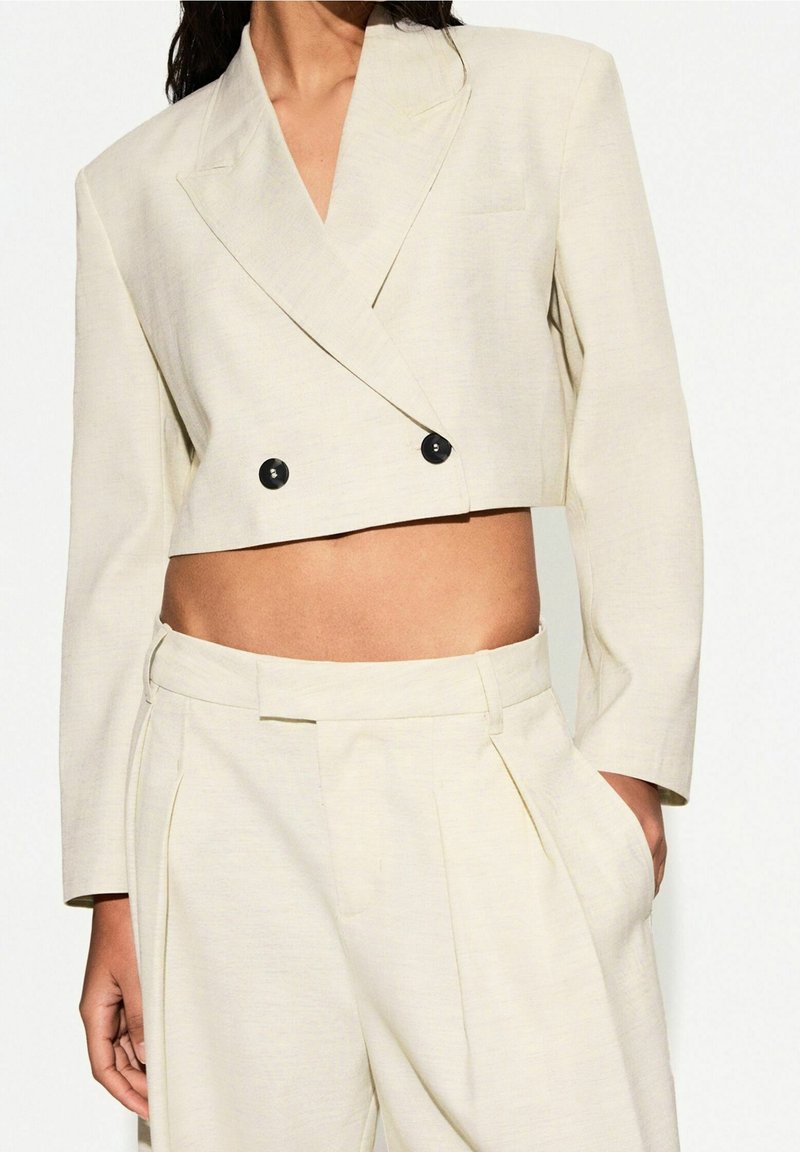 Femme portant un blazer court crème à double boutonnage avec des boutons noirs et un pantalon plissé taille haute, main dans la poche.