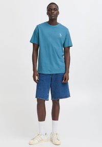 Teal bomulds T-shirt med korte ærmer, parret med knælange blå denimshorts. Hvide sneakers og hvide sokker i crew-længde fuldender looket.