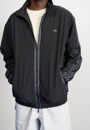 Mann trägt eine schwarze Adidas-Jacke mit Reißverschluss und drei reflektierenden Streifen an den Ärmeln sowie weiße Shorts mit Taschen.