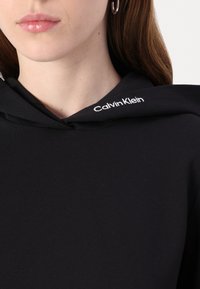 Crna dukserica s kapuljačom od mekanog materijala, s bijelim logotipom "Calvin Klein" na kapuljači, detaljni prikaz s naglaskom na izrez i tkaninu.