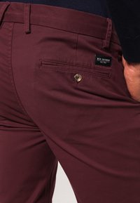 Bordeauxrode broek van een gladde stof met een knoopsluiting en een gestikte achterzak met een zwart label.