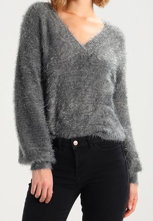 Femme portant un pull gris duveteux à col en V associé à un jean noir taille haute, debout devant un fond clair uni.
