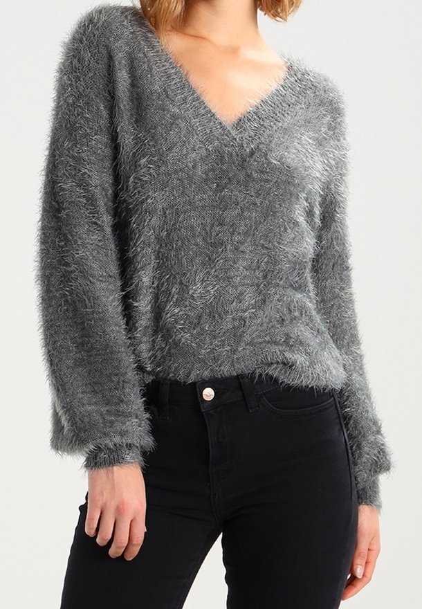 Femme portant un pull gris duveteux à col en V associé à un jean noir taille haute, debout devant un fond clair uni.