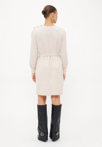 Beige gebreide jurk met lange mouwen, een getailleerde taille en knoopdetails op de schouders, gecombineerd met zwartleren laarzen tot de knie.