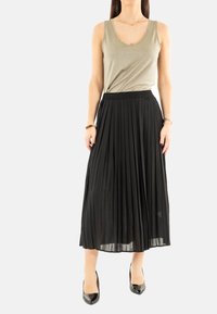 Jupe maxi plissée noire avec une taille élastique, associée à un débardeur scintillant olive. Équipée de talons plats et pointus noirs.