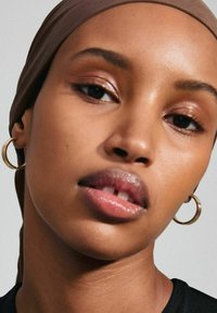 Pendientes de aro dorados en una persona con piel marrón suave, que lleva un pañuelo marrón y una blusa negra, con una iluminación suave que resalta las características faciales.