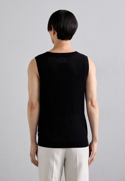 Black sleeveless knit top with a ribbed texture, featuring side slits, worn over light-colored trousers. The back shows a clean, minimalist design.