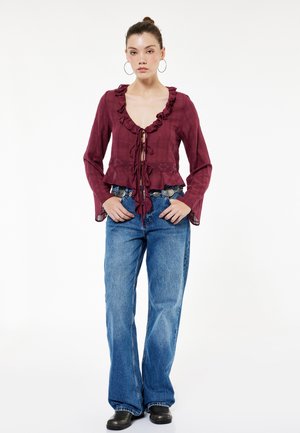 Vrouw die een bordeauxrode blouse met ruches en strik voor draagt, wijd uitlopende blauwe jeans, decoratieve riem, zwarte schoenen en grote oorringen, staand.