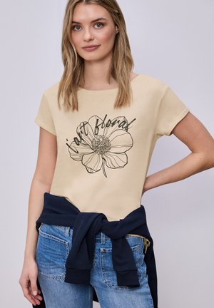 Jeune femme portant un t-shirt beige avec un motif floral linéaire noir et du texte, un jean bleu et un sweat-shirt bleu marine noué autour de la taille.