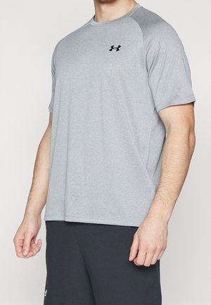 Mann trägt hellgraues kurzärmeliges Under Armour Sportshirt und dunkelgraue Shorts, steht vor weißem Hintergrund.
