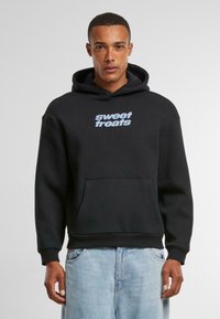 Black hoodie made of cotton with a front pocket, featuring "sweet treats" in light blue text on the chest. Classic fit and textured fabric.