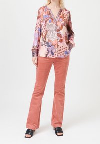 Blouse à motifs floraux aux tons roses, violets et bleus avec un décolleté en V ; associée à un pantalon évasé en velours côtelé corail et des sandales à talons noires.