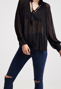 Blusa nera trasparente con un design plissettato, colletto con fiocco e maniche lunghe. Indossata con jeans skinny scuri strappati.