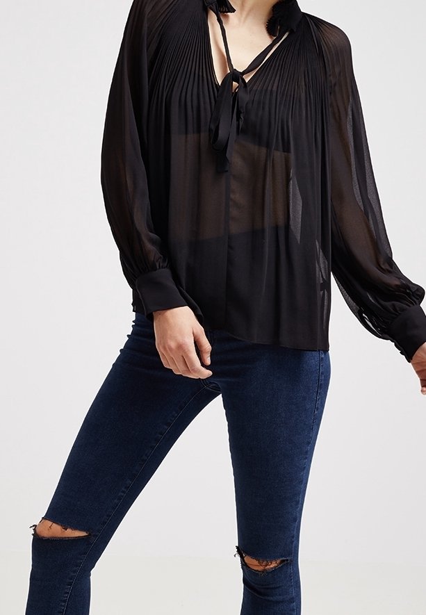 Blusa nera trasparente con un design plissettato, colletto con fiocco e maniche lunghe. Indossata con jeans skinny scuri strappati.