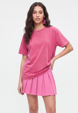 Femme aux longs cheveux foncés portant un t-shirt rose délavé oversize, une jupe plissée rose vif et des boucles d'oreilles roses en forme d'étoile, posant avec une main sur la hanche.