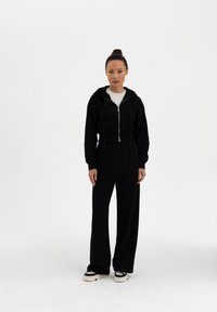 Mustapohja crop-huppari, jossa on naru ja vetoketju, pitkät hihat, yhdistetty korkeavyötäröisiin mustiin housuihin, jotka ovat rento mitoitukseltaan ja leveitä lahkeita.