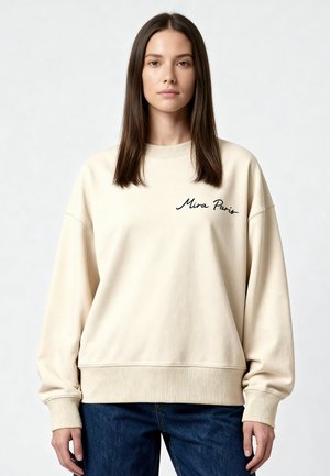 Giovane donna con capelli castani lisci indossa una felpa crema oversize con la scritta "Mira Paris", abbinata a jeans blu, in piedi su sfondo bianco.