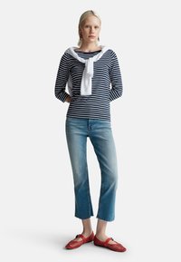 Granatowo-białe paskowane bluzka z długim rękawem, z białym swetrem związanym na szyi, w połączeniu z jasnoniebieskimi dżinsami 7/8 i czerwonymi perforowanymi balerinami.