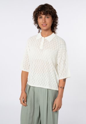 Frau mit lockigem dunklem Haar, die ein weißes gestricktes Polo-Shirt mit kurzen Ärmeln und lockere hellgrüne Hosen trägt, steht vor einem einfarbigen Hintergrund.